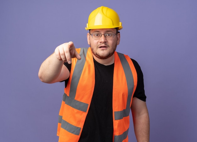 Uomo del costruttore in giubbotto da costruzione e casco di sicurezza che punta con il dito indice alla telecamera guardando confuso in piedi sopra il blu