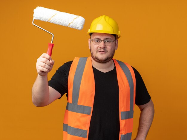 Uomo del costruttore in giubbotto da costruzione e casco di sicurezza che tiene il rullo di vernice che guarda la telecamera