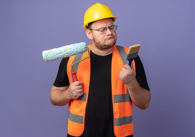 Uomo del costruttore in giubbotto da costruzione e casco di sicurezza che tiene rullo di vernice e pennello che sembra confuso cercando di fare una scelta in piedi su sfondo blu