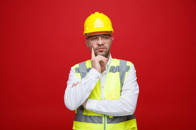 Uomo del costruttore in uniforme da costruzione e casco di sicurezza che indossa occhiali di sicurezza che guarda da parte con un'espressione pensierosa che pensa in piedi su sfondo rosa