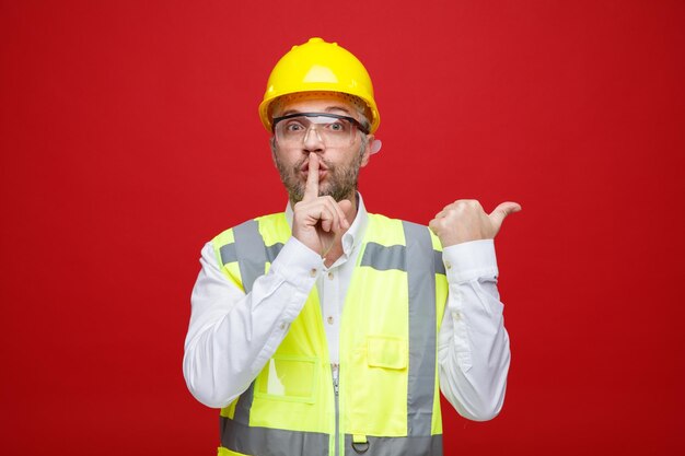 Uomo del costruttore in uniforme da costruzione e casco di sicurezza che indossa occhiali di sicurezza guardando la fotocamera facendo un gesto di silenzio con il dito sulle labbra che punta con il pollice di lato in piedi su sfondo rosso