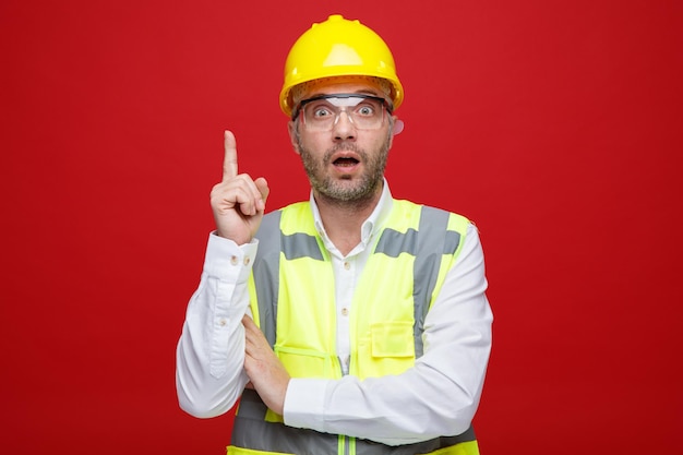 Uomo del costruttore in uniforme da costruzione e casco di sicurezza che indossa occhiali di sicurezza guardando la fotocamera sorpreso e stupito che mostra il dito indice in piedi su sfondo rosa