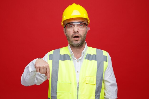 Uomo del costruttore in uniforme da costruzione e casco di sicurezza che indossa occhiali di sicurezza guardando la fotocamera sorpreso puntando con il dito indice verso il basso in piedi su sfondo rosa