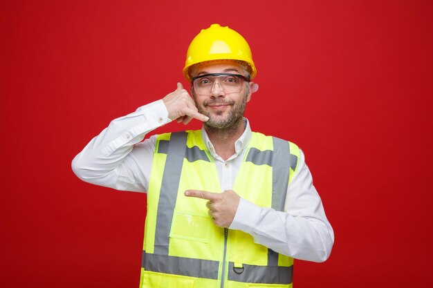 Uomo del costruttore in uniforme da costruzione e casco di sicurezza che indossa occhiali di sicurezza guardando la fotocamera sorridente amichevole facendomi chiamare gesto puntando con il dito indice di lato in piedi su sfondo rosso