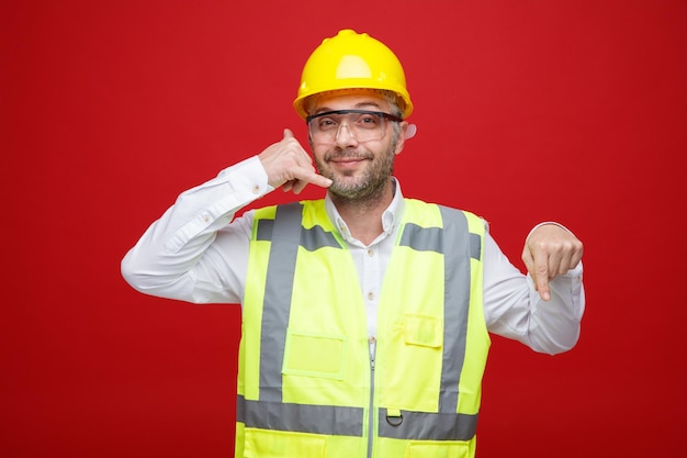 Uomo del costruttore in uniforme da costruzione e casco di sicurezza che indossa occhiali di sicurezza guardando la fotocamera sorridente amichevole facendomi chiamare gesto puntando con il dito indice verso il basso in piedi su sfondo rosso