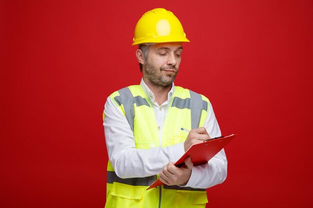 Uomo del costruttore in uniforme da costruzione e casco di sicurezza che tiene appunti che sembra sicuro di prendere appunti in piedi su sfondo rosso