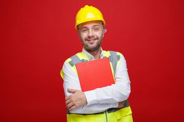 Uomo del costruttore in uniforme da costruzione e casco di sicurezza che tiene appunti guardando la fotocamera sorridente felice e positivo in piedi su sfondo rosso