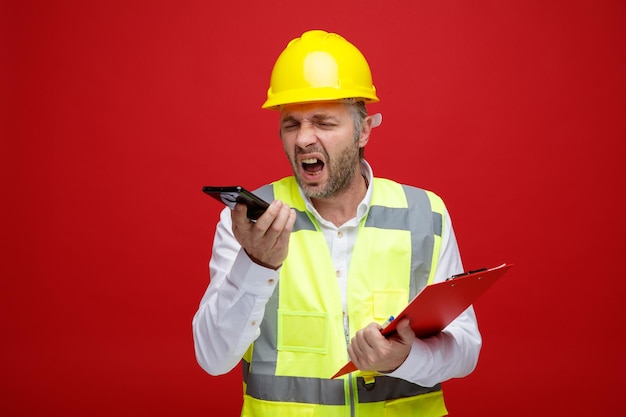 Uomo del costruttore in uniforme da costruzione e casco di sicurezza che tiene appunti parlando sul telefono cellulare impazzendo gridando con espressione aggressiva in piedi su sfondo rosso