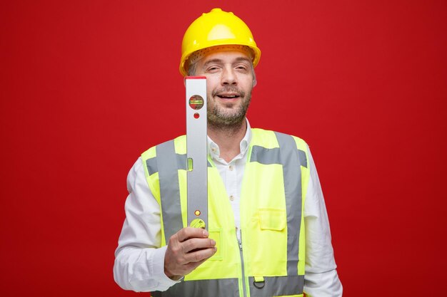 Uomo del costruttore in uniforme da costruzione e casco di sicurezza che tiene il righello di livello guardando la fotocamera con un grande sorriso sulla faccia felice in piedi su sfondo rosso