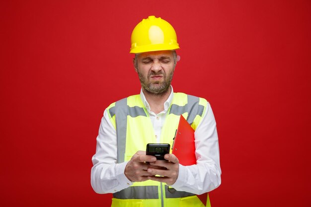 Uomo del costruttore in uniforme da costruzione e casco di sicurezza che tiene un messaggio di testo negli appunti utilizzando lo smartphone accigliato facendo la bocca storta essendo insoddisfatto in piedi su sfondo rosso