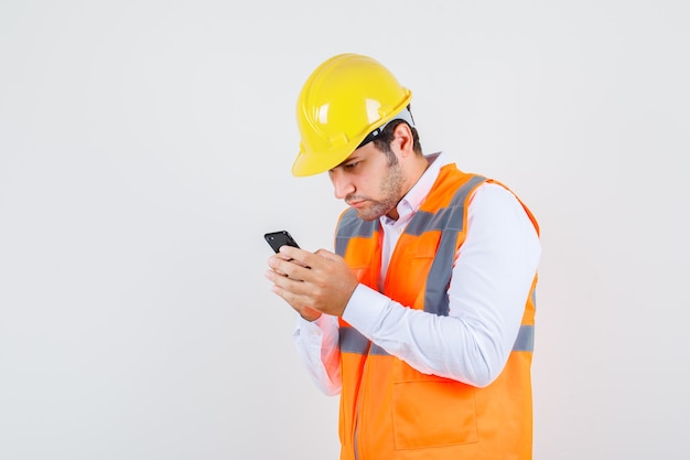 Uomo del costruttore utilizza lo smartphone in camicia, uniforme e sembra occupato. vista frontale.