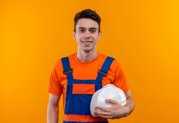 Uomo del giovane costruttore che indossa l'uniforme della costruzione che sorride e che tiene il casco di sicurezza