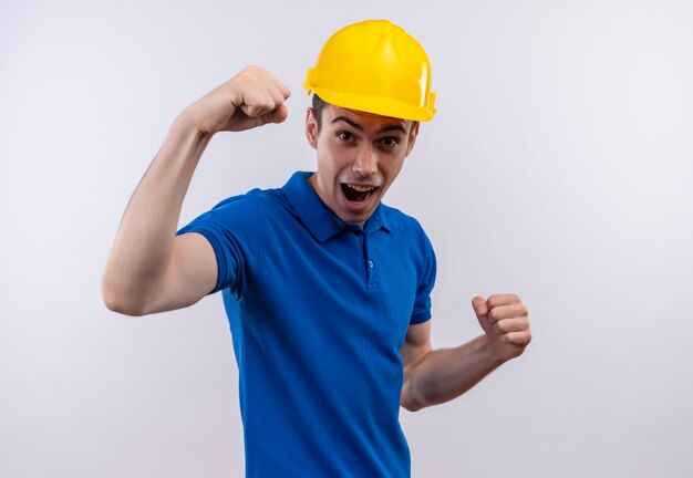 Uomo del giovane costruttore che indossa l'uniforme della costruzione e il casco di sicurezza che fa fronte felice e aumentano i pugni