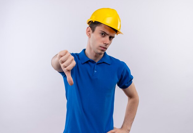 Uomo del giovane costruttore che indossa l'uniforme della costruzione e il casco di sicurezza che fa i pollici arrabbiati giù