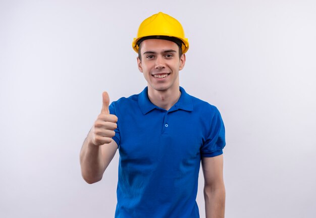 Uomo del giovane costruttore che indossa l'uniforme della costruzione e il casco di sicurezza che fa i pollici felici in su