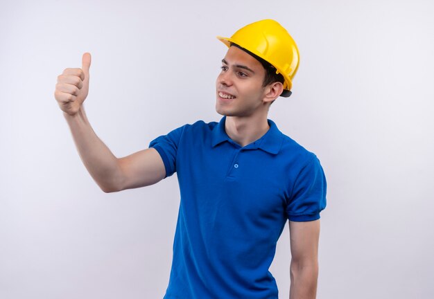 Uomo del giovane costruttore che indossa l'uniforme della costruzione e il casco di sicurezza che fa i pollici felici in su