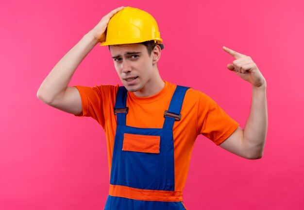 Uomo del giovane costruttore che indossa l'uniforme della costruzione e il punto del casco di sicurezza con il pollice sul casco