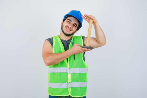 Uomo del giovane costruttore che mostra le dimensioni del martello in uniforme da lavoro e che sembra allegro, vista frontale.