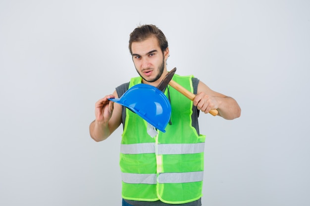 Uomo del giovane costruttore che tiene casco e martello in uniforme da lavoro e che sembra fiducioso. vista frontale.