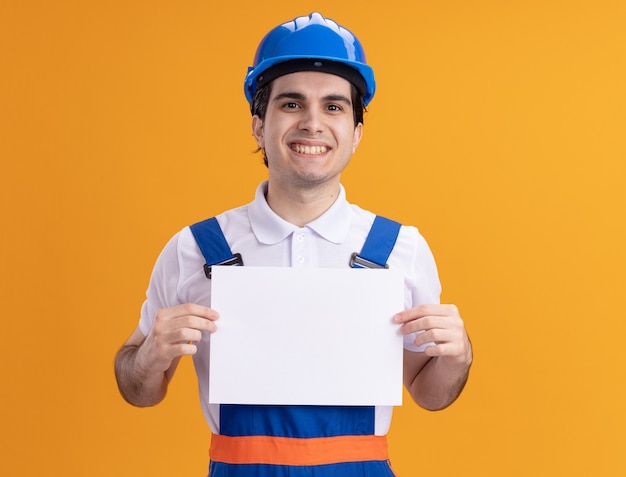 Uomo del giovane costruttore in uniforme da costruzione e casco di sicurezza che tiene la pagina vuota guardando la parte anteriore sorridente allegramente in piedi sopra la parete arancione