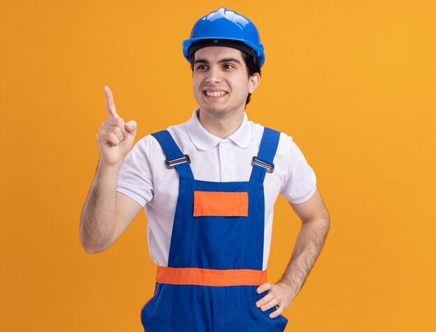 Uomo del giovane costruttore in uniforme della costruzione e casco di sicurezza che osserva da parte sorridente fiducioso che indica con il dito indice a qualcosa che si leva in piedi sopra la parete arancione