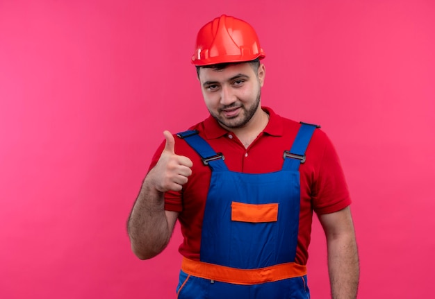 Uomo del giovane costruttore in uniforme della costruzione e casco di sicurezza che sorride allegramente mostrando i pollici in su