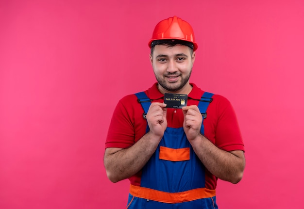 Uomo del giovane costruttore in uniforme della costruzione e casco di sicurezza che tiene la carta di credito che sorride con la faccia felice