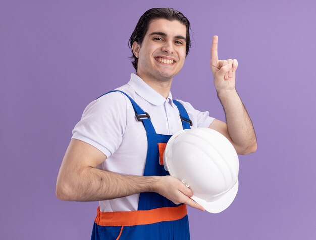 Uomo del giovane costruttore in uniforme di costruzione che tiene il suo casco di sicurezza guardando davanti con espressione fiduciosa che punta con il dito indice in alto sorridente in piedi sopra la parete viola