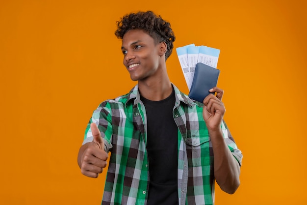 Uomo del giovane viaggiatore afroamericano che tiene i biglietti aerei che sorride allegramente, positivo e felice che osserva da parte che mostra i pollici in su