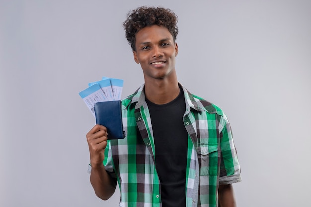 Uomo del giovane viaggiatore afroamericano che tiene i biglietti aerei che sorride allegramente positivo e felice guardando la fotocamera in piedi su sfondo bianco