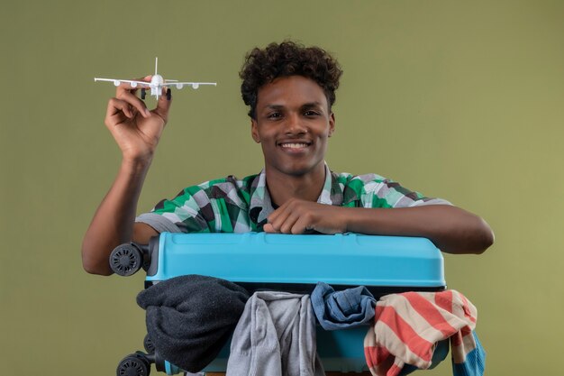 Uomo del giovane viaggiatore afroamericano in piedi con la valigia piena di vestiti che tiene aereo giocattolo guardando la telecamera sorridendo allegramente su sfondo verde