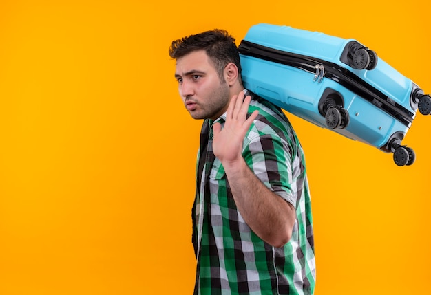 Uomo del giovane viaggiatore in camicia controllata che tiene la valigia che osserva da parte con la faccia seria che fluttua con la mano che sta sopra la parete arancione