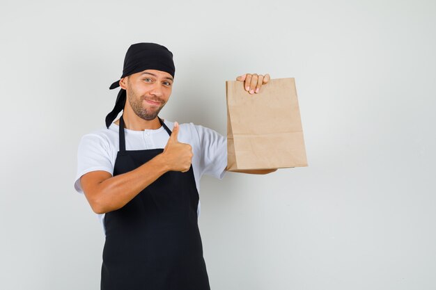 Uomo del panettiere che tiene il sacchetto di carta, mostrando il pollice in su in maglietta