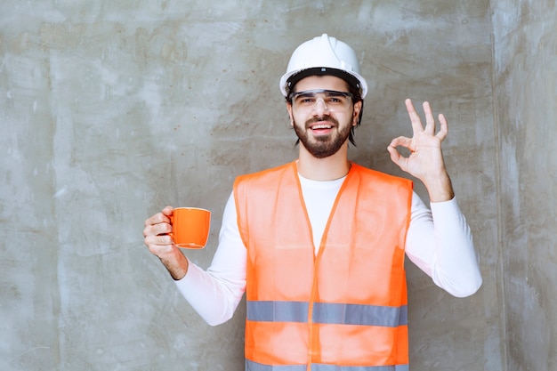 Uomo dell'ingegnere in casco bianco che tiene una tazza arancione e che gode del gusto della bevanda.