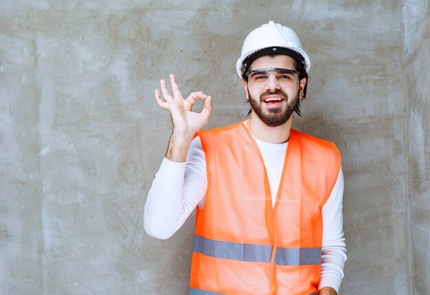 Uomo dell'ingegnere in casco bianco e occhiali protettivi che mostrano il segno giusto della mano.