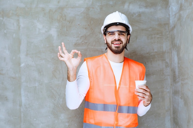 Uomo dell'ingegnere in casco bianco e occhiali protettivi che tengono una tazza di bevanda e che mostrano il segno di godimento.