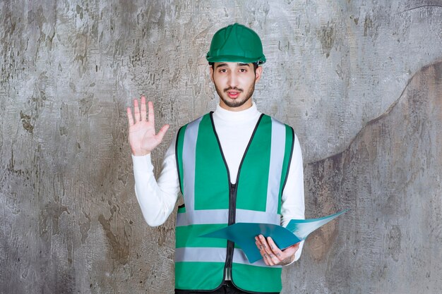 Uomo dell'ingegnere in uniforme gialla e casco che tiene una cartella blu e che ferma qualcosa.