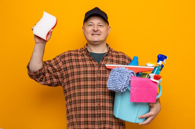 Uomo delle pulizie in camicia a quadri e cappello che tiene il secchio con strumenti per la pulizia e spugna che guarda con un sorriso sul viso