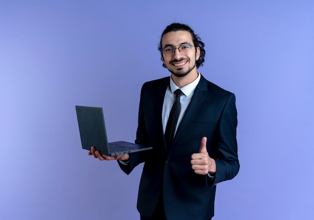 Uomo di affari in vestito nero e occhiali che tengono computer portatile che guarda al fronte sorridente che mostra i pollici in su che si leva in piedi sopra la parete blu