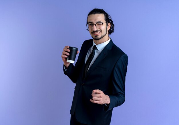 Uomo di affari in vestito nero e vetri che tengono il pugno di serraggio della tazza di caffè felice e positivo sorridente e ammiccante che sta sopra la parete blu