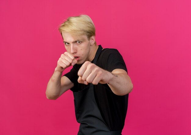 Uomo di barbiere professionista in grembiule che punta con il pugno chiuso alla macchina fotografica con la faccia seria