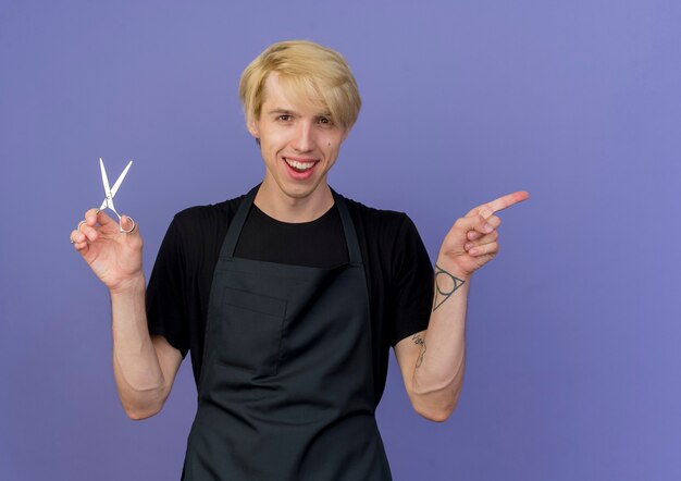 Uomo di barbiere professionista in grembiule che tiene le forbici pointin con il dito indice al lato sorridente con la faccia felice