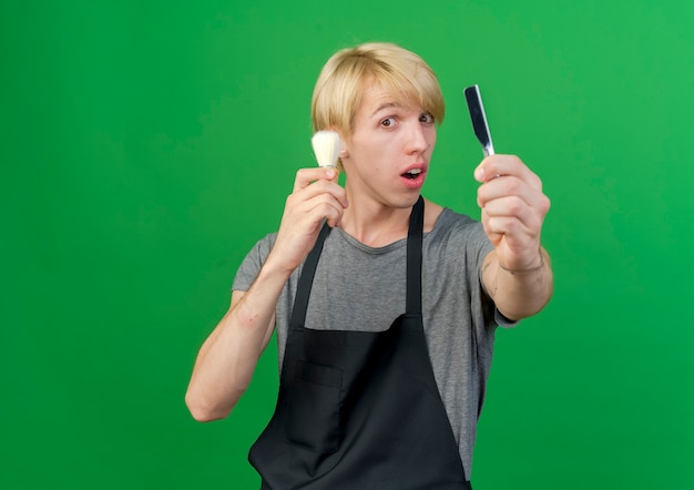 Uomo di barbiere professionista in grembiule che tiene rasoio e pennello da barba che sembra sorpreso