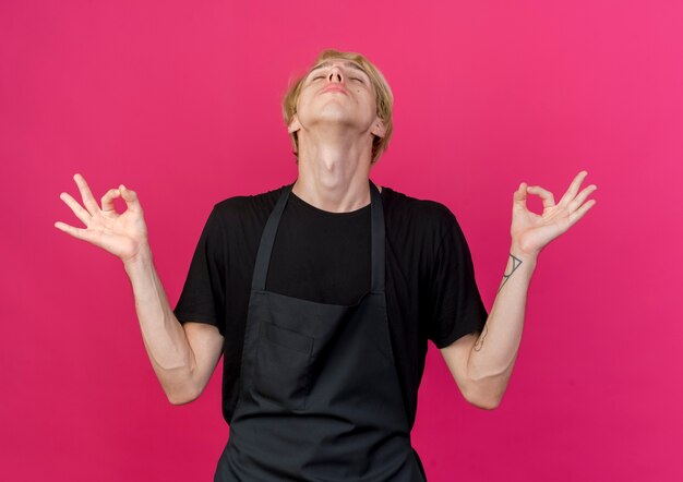 Uomo di barbiere professionista in grembiule rilassante con gli occhi chiusi che fa il gesto di meditazione con le dita