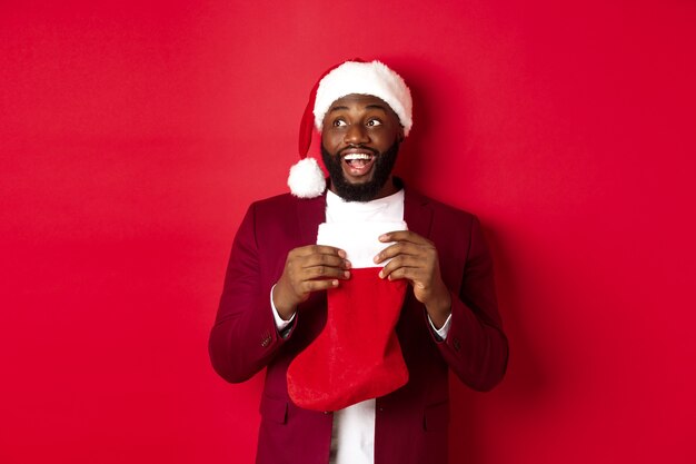 Uomo di colore allegro che guarda l'angolo in alto a sinistra e sorride, tiene in mano un calzino di Natale con regali, in piedi su uno sfondo rosso