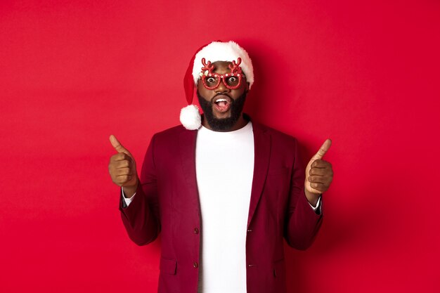 Uomo di colore divertente che celebra il nuovo anno, indossando occhiali da festa e cappello da Babbo Natale, mostrando il pollice in su, approva e piace, in piedi su sfondo rosso.