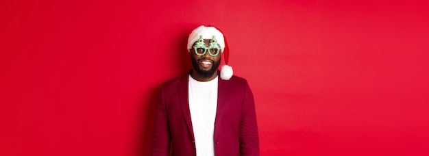 Uomo di colore divertente in cappello di Babbo Natale e occhiali da festa che celebrano il Natale sorridendo felice e augurando buon