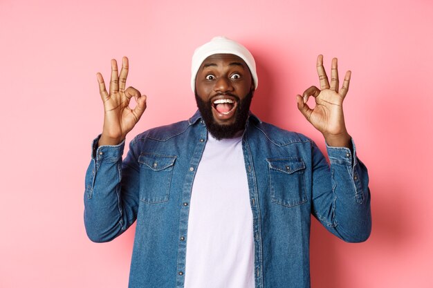 Uomo di colore impressionato in berretto e camicia, mostrando segni ok e fissando la telecamera stupito, sorridendo felice, lodando la grande offerta, consigliando il prodotto, sfondo rosa