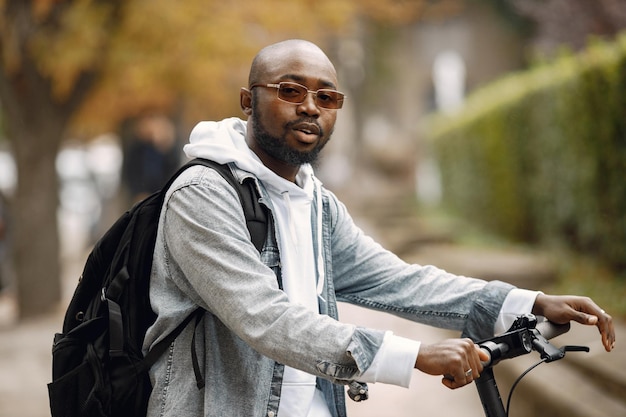 Uomo di colore in piedi su una strada con scooter elettrico. Uomo che indossa una felpa con cappuccio bianca e jeans neri