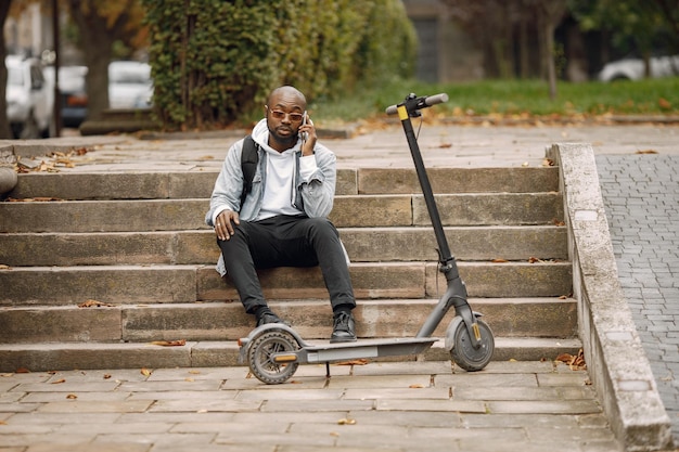 Uomo di colore seduto su una strada con scooter elettrico. Uomo che indossa una felpa con cappuccio bianca e jeans neri